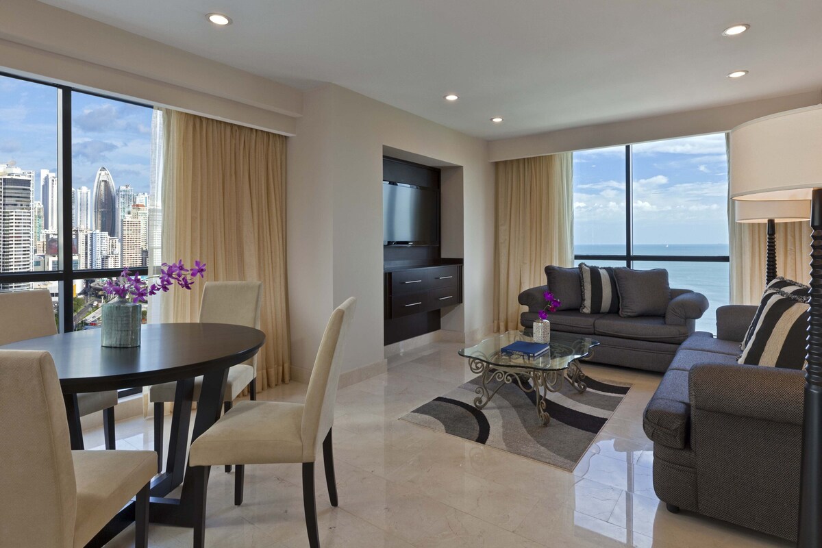 A modern living room with a dining area, large windows, a flat-screen TV, and a city view.