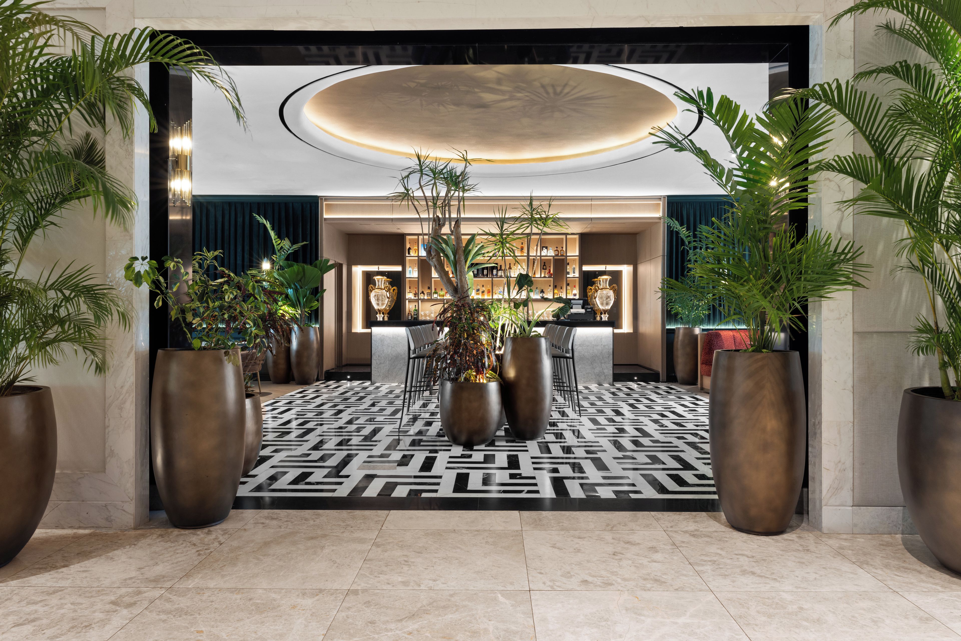 Un vestíbulo de hotel con piso estampado, grandes plantas en macetas, una zona de bar y un mostrador de recepción.
