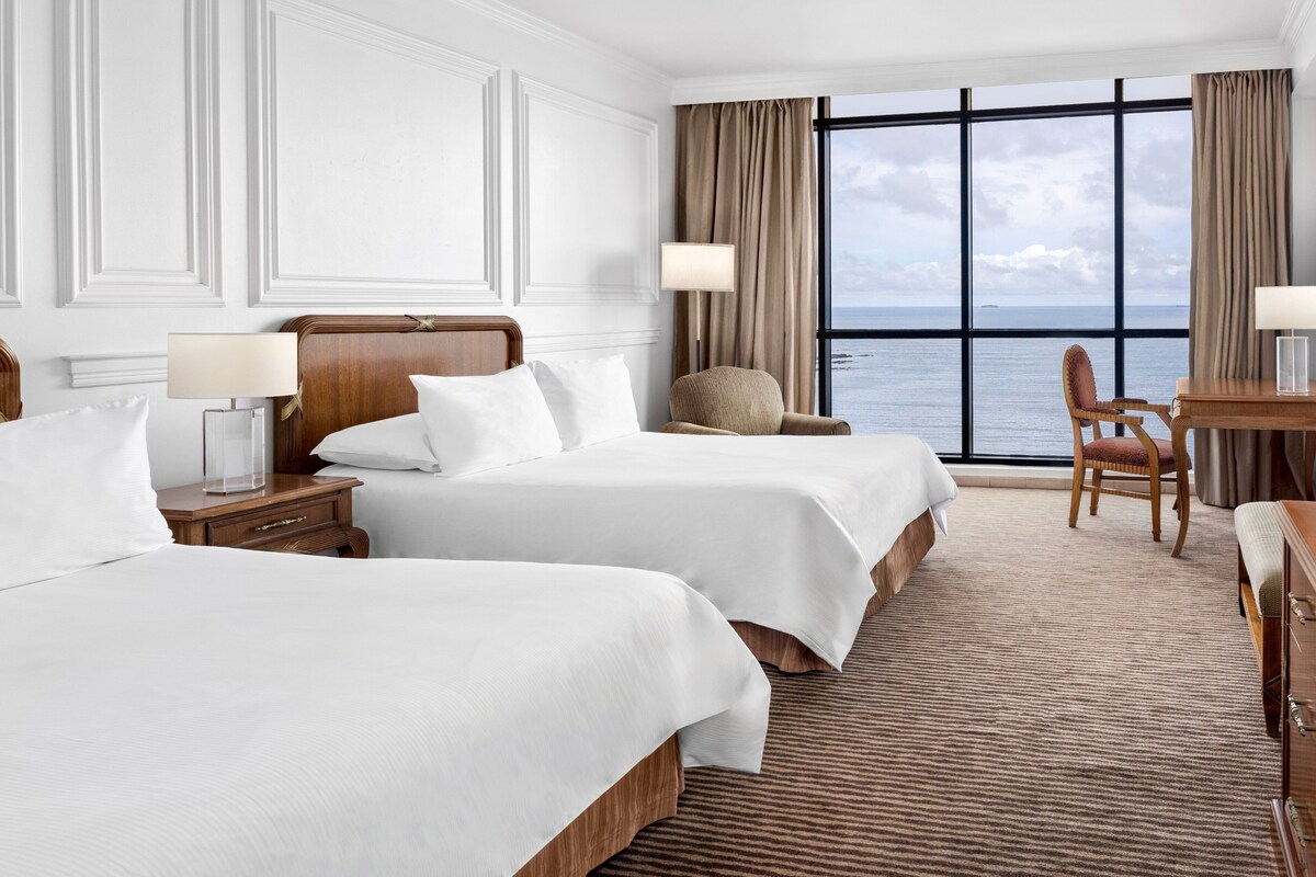 A hotel room with two beds, a desk, a chair, and a large window with a view of the sea.