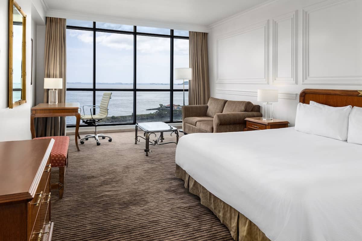 A hotel room with a large bed, a desk, a chair, and a view of the sea.
