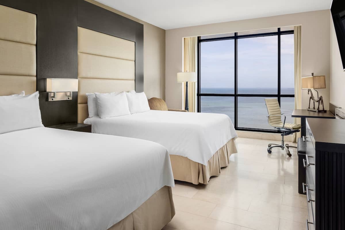 A hotel room with two beds, a desk, a chair, a lamp, and a view of the ocean.