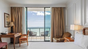 Quarto de hotel com cama, escrivaninha, cadeira, mesa de cabeceira e vista para o oceano.