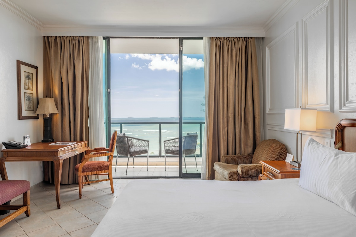 Una habitación de hotel con cama, escritorio, silla, mesita de noche y vista al océano.
