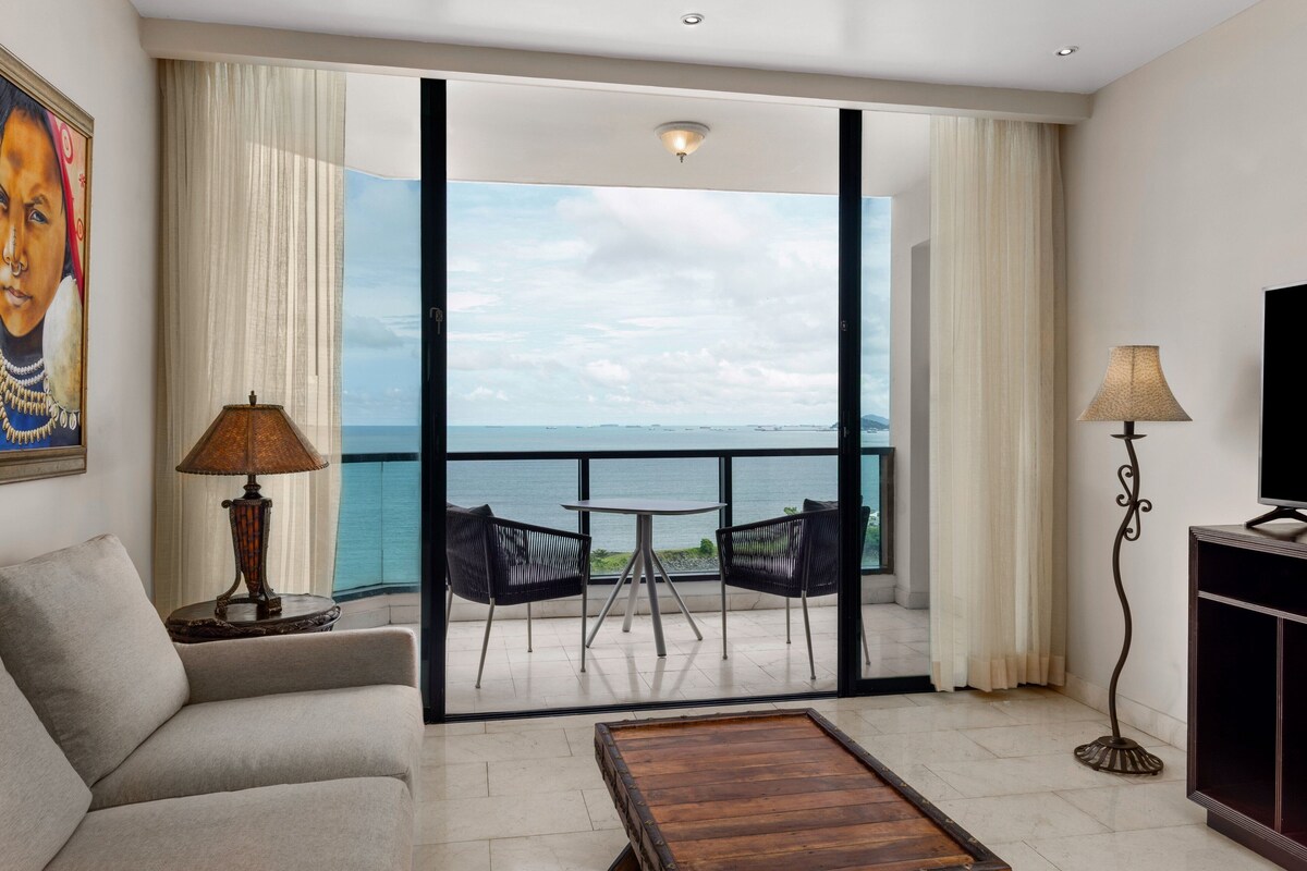 A room with a sofa, a coffee table, a TV stand, a lamp, and a view of the sea through large windows.
