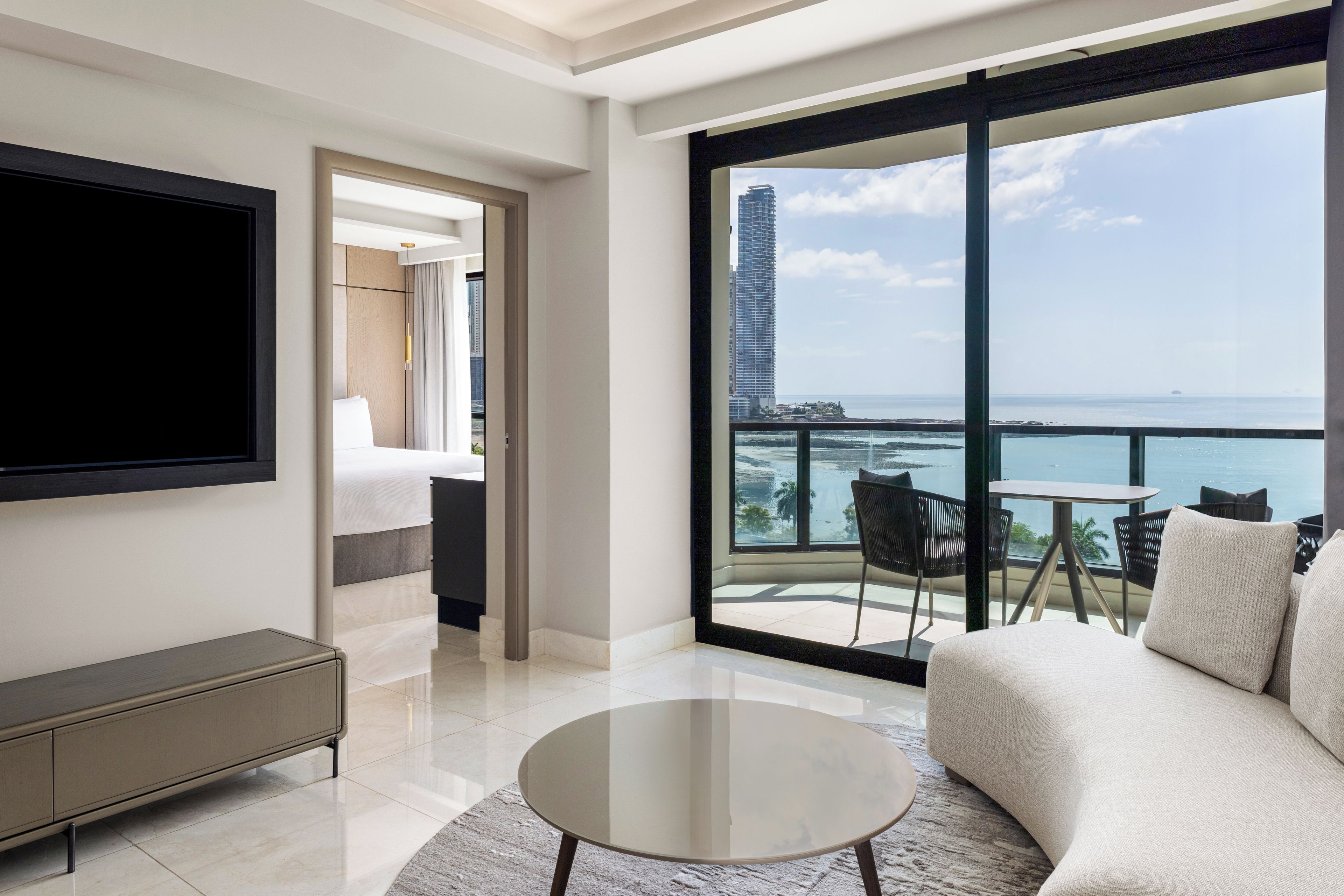 Habitación de hotel moderna con un balcón grande, televisor de pantalla plana, sofá y mesa de comedor.
