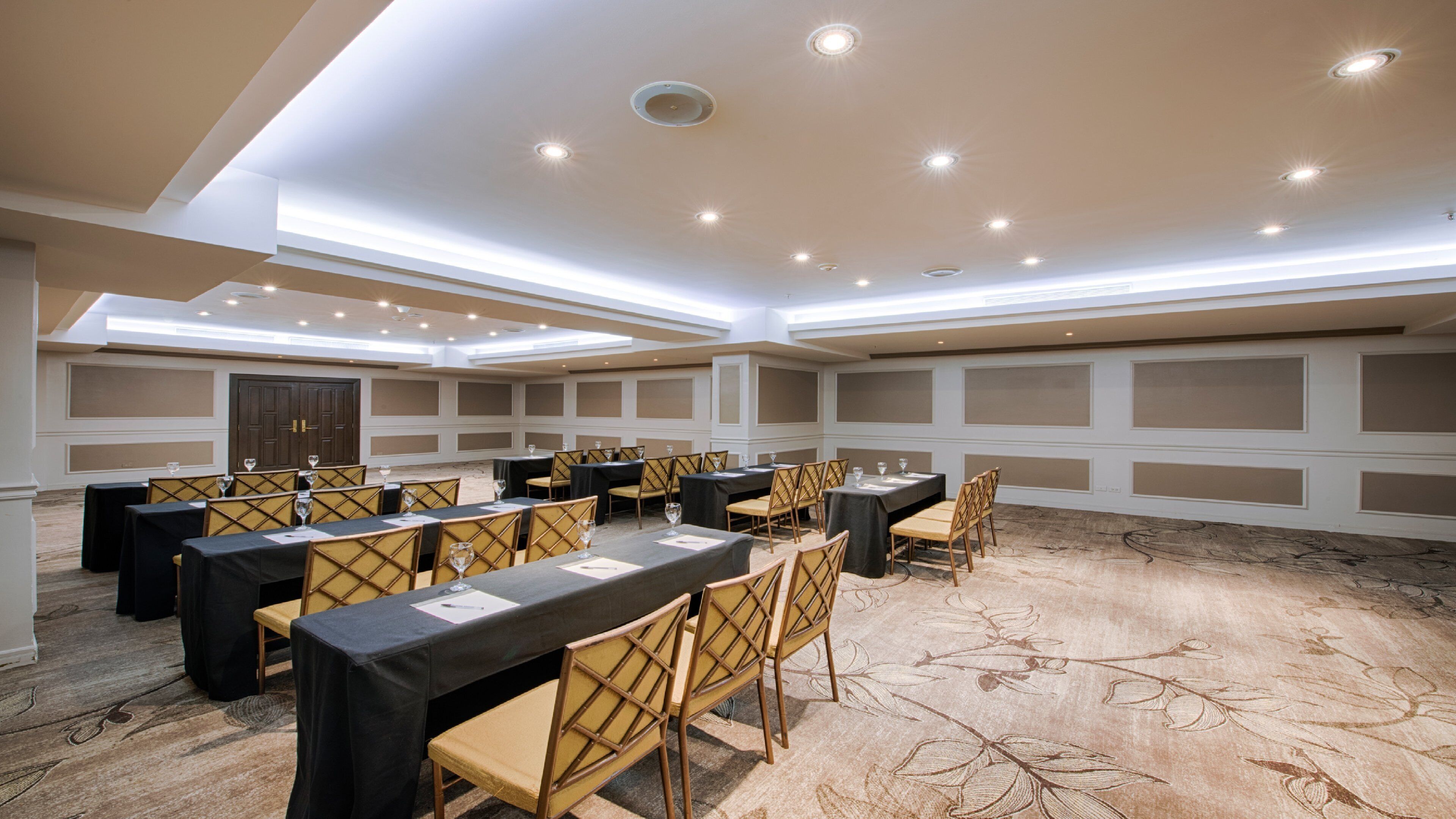 Un salón de conferencias con varias mesas y sillas, iluminación empotrada y alfombra estampada.