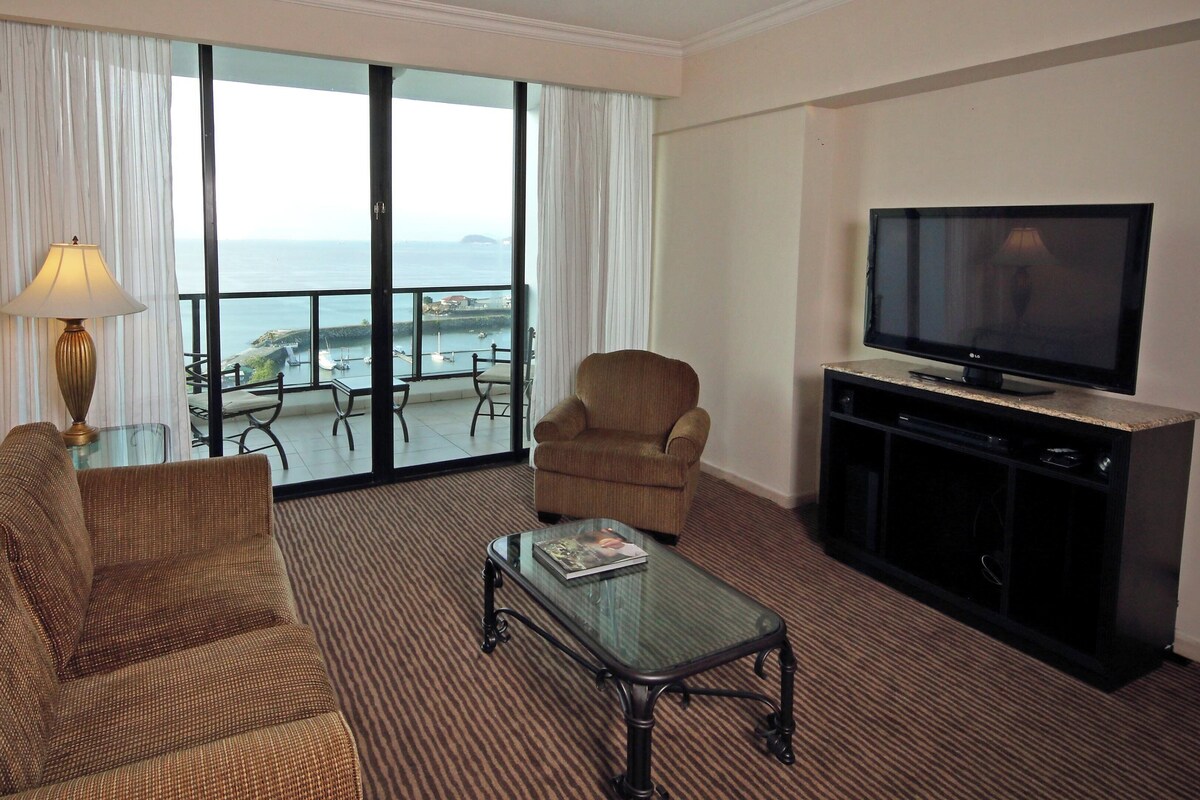 A hotel room with a sofa, armchair, glass coffee table, TV, and a balcony with a view of the sea.