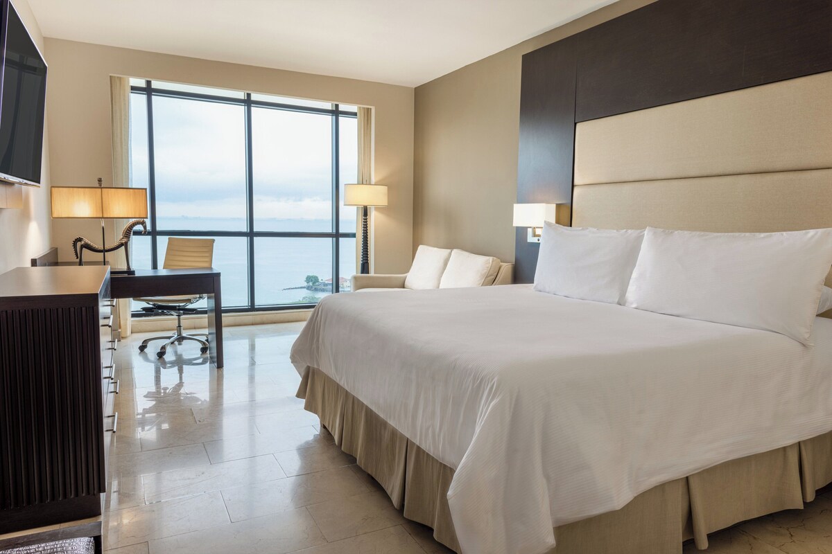 A hotel room with a large bed, two bedside tables, a desk with a chair, and a large window offering a view.