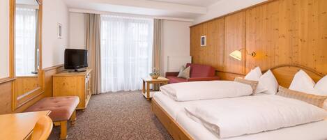 Una habitación de hotel con cabecera de madera, cama doble, escritorio con televisor, una mesita con una planta y una silla roja.