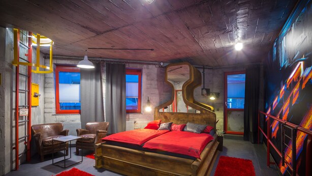 A bedroom with a wooden bed, red bedding, a red rug, a desk with a chair, and a window with blinds. - Kongress Hotel (Leoben)