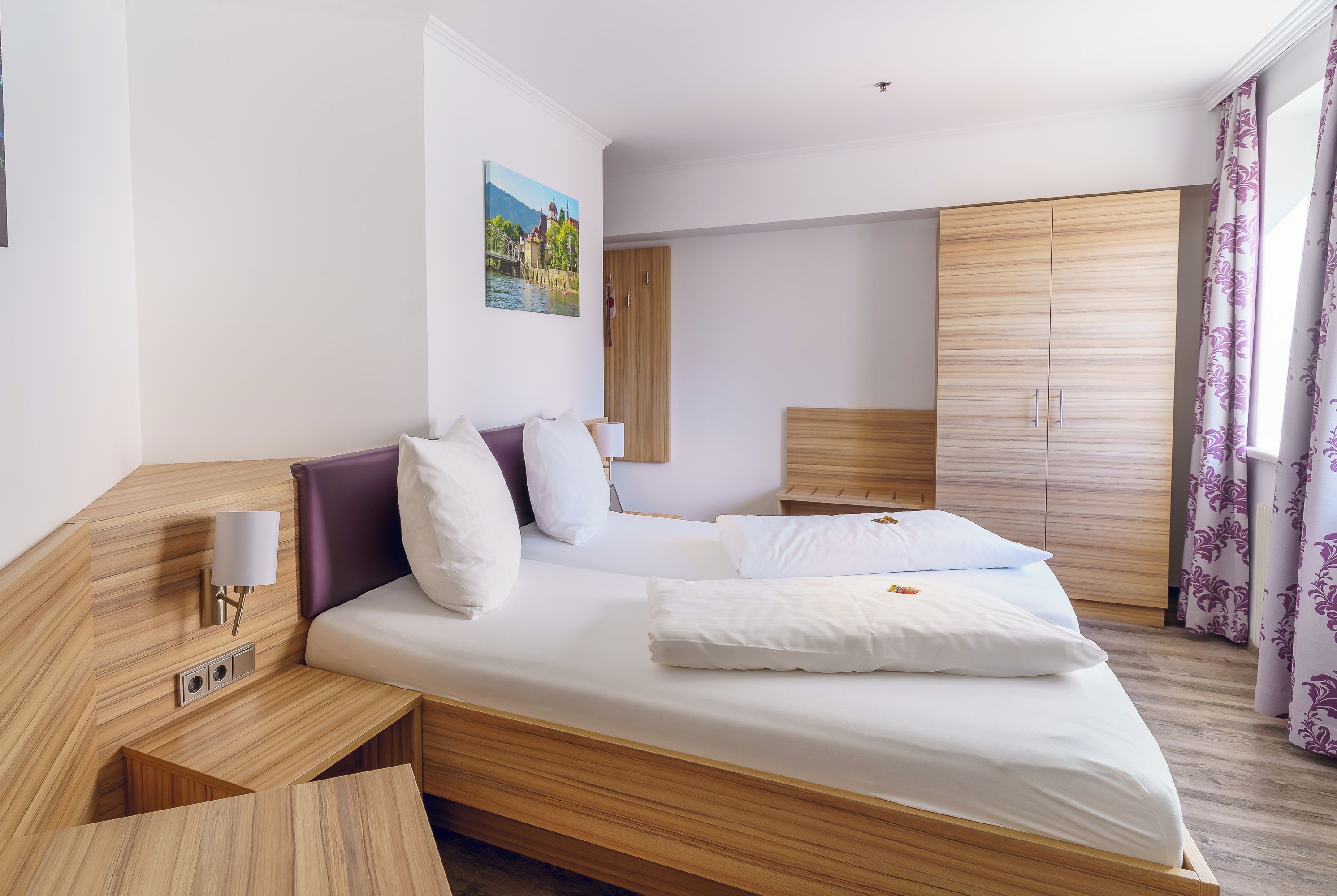A modern hotel room with a wooden bed, bedside tables, a nightstand, a wardrobe, and a painting on the wall.