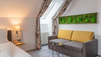 Una habitación de hotel moderna con una cama, un sofá con cojines amarillos, una mesita redonda pequeña, una lámpara de pared y un cuadro de arte en la pared con musgo verde.