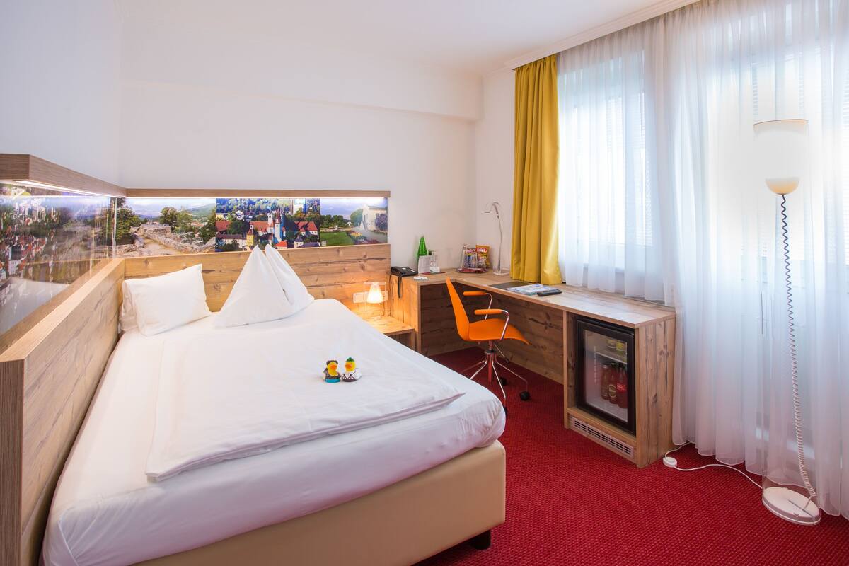 A hotel room with a bed, a desk, a chair, a lamp, and a view of a cityscape through a window.