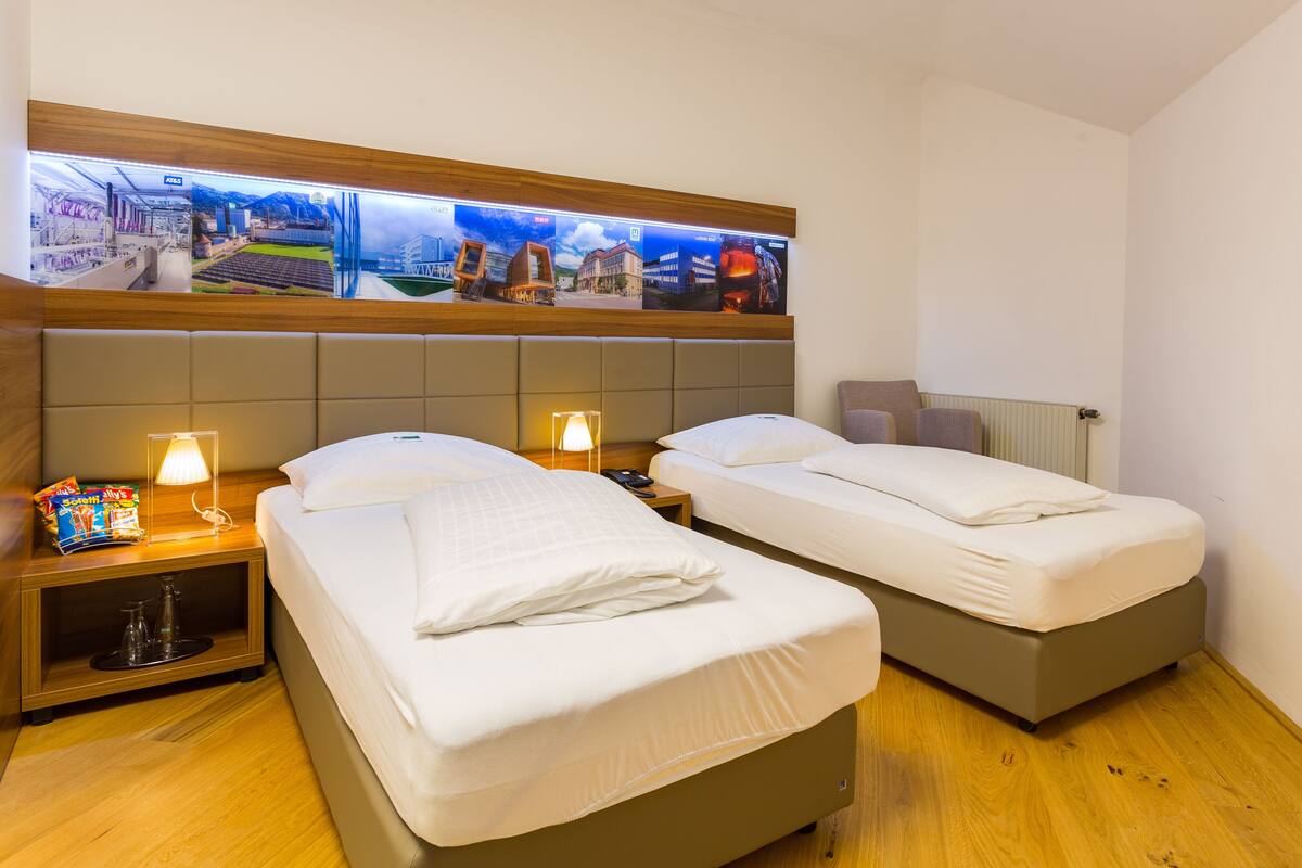 A hotel room with two beds, a wooden headboard, a wall-mounted TV displaying cityscapes, and a small table with a lamp and snacks.
