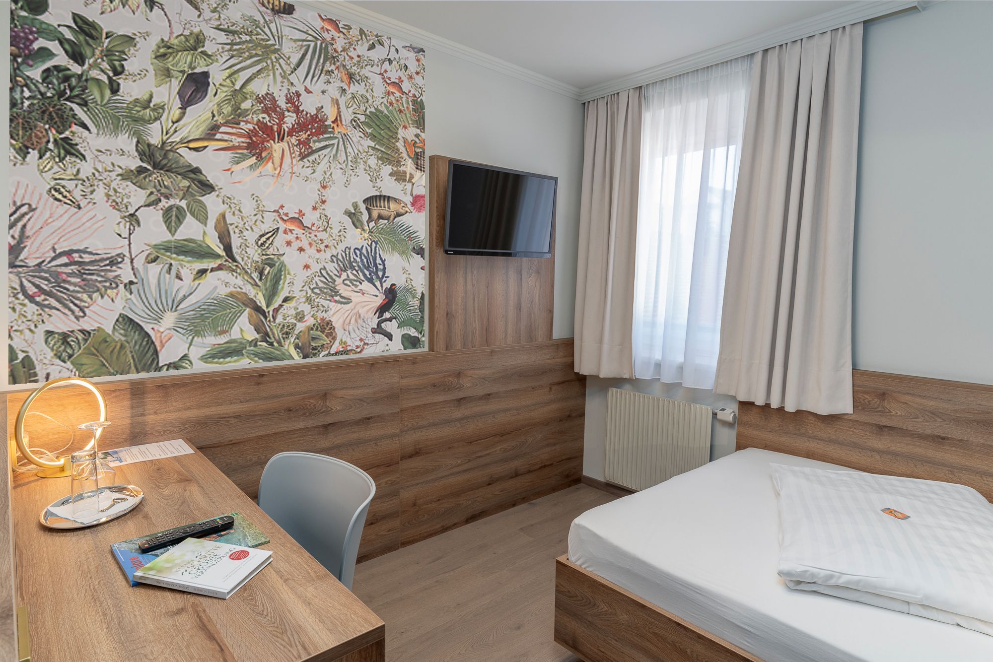 A hotel room with a wooden headboard, a bed, a desk with a chair, a television mounted on the wall, and a window with curtains.