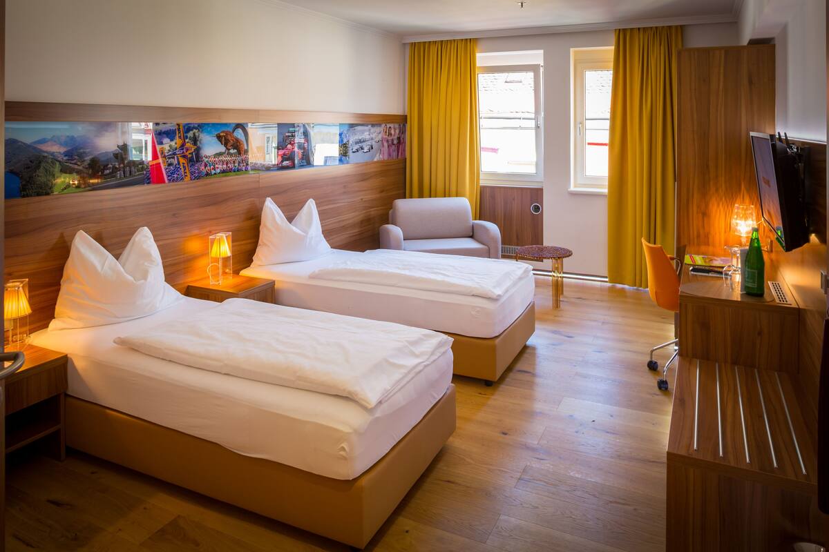 A hotel room with two beds, a wooden headboard, a small table, a chair, a TV, and a painting on the wall.