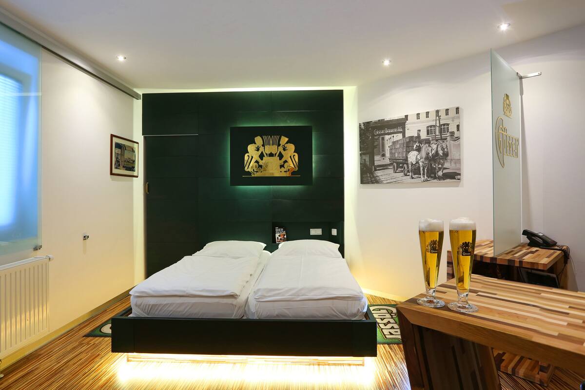 A modern hotel room with a bed, a wooden side table with two glasses of beer, and a framed picture on the wall.