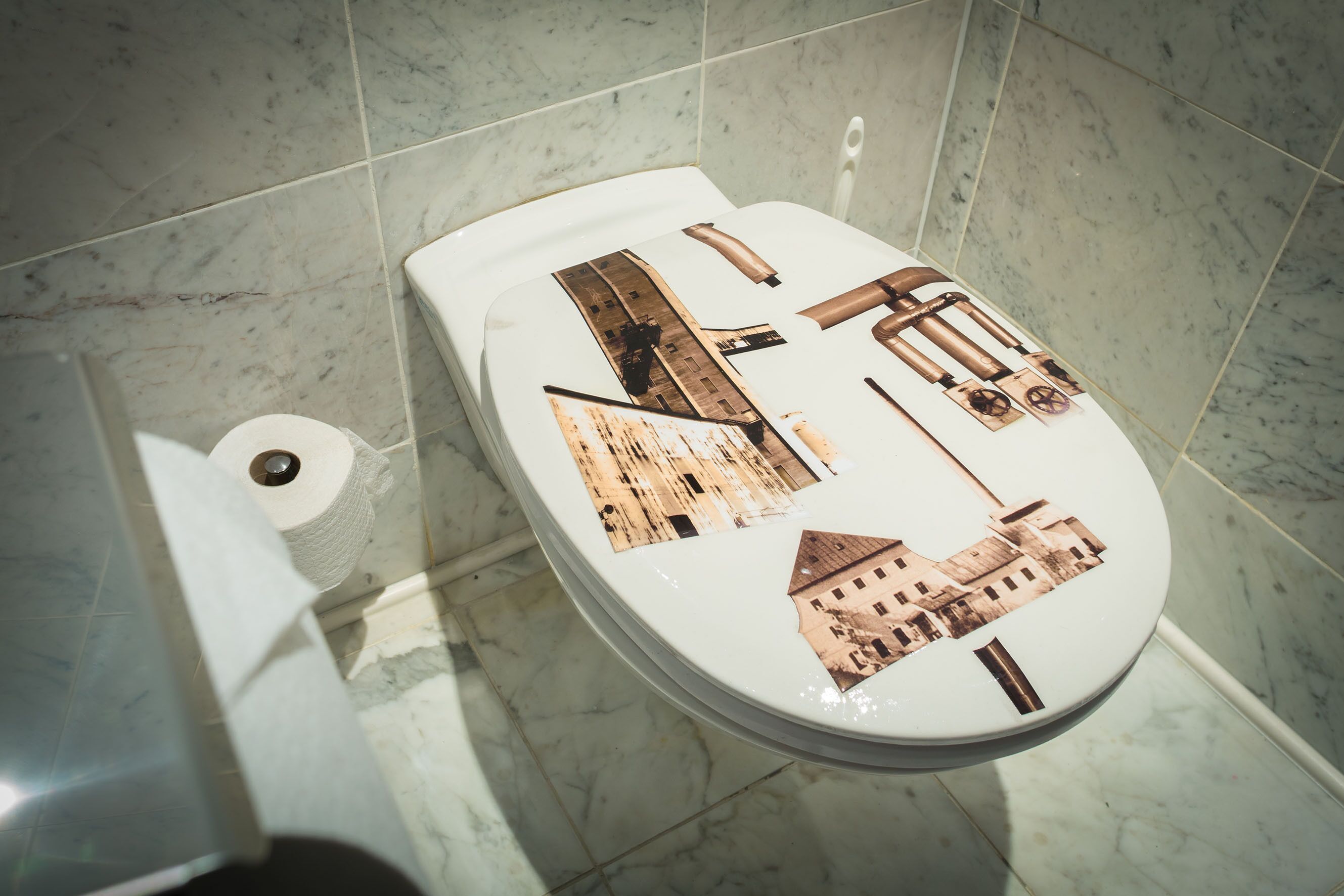 Un inodoro con piso y paredes de mármol, un rollo de papel higiénico y una palanca para accionar la descarga.