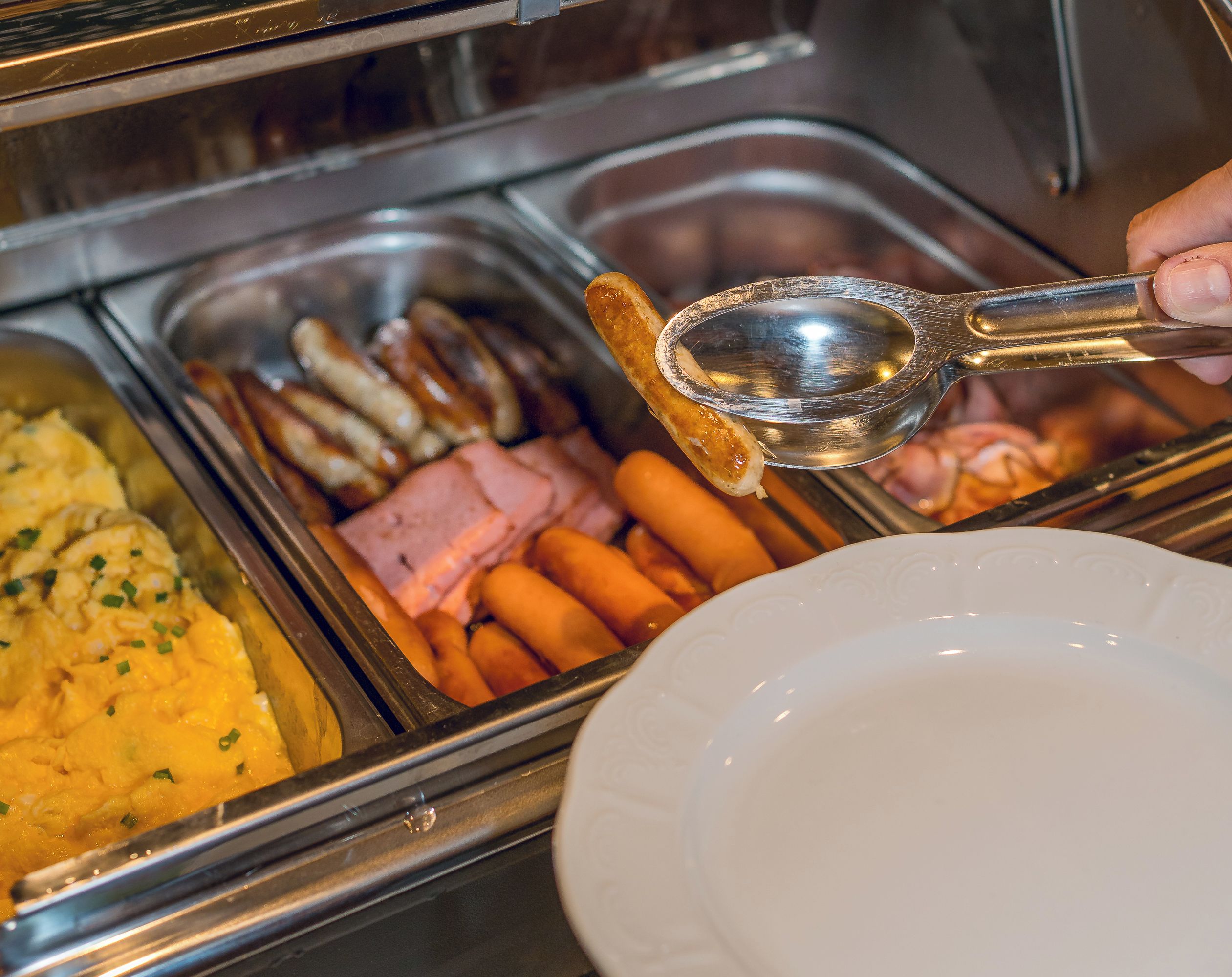 A buffet with various food items including sausages, ham, and scrambled eggs.