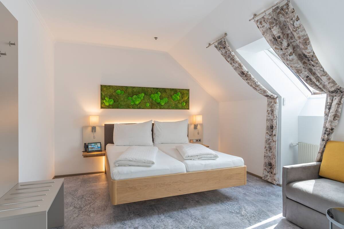A modern bedroom with a wooden bed, a green wall art piece, a nightstand with a lamp, and a view of the outdoors through a window with curtains.