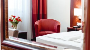 Una habitación de hotel con una cama, un sillón rojo, una mesita de noche con un teléfono y un jarrón con flores.