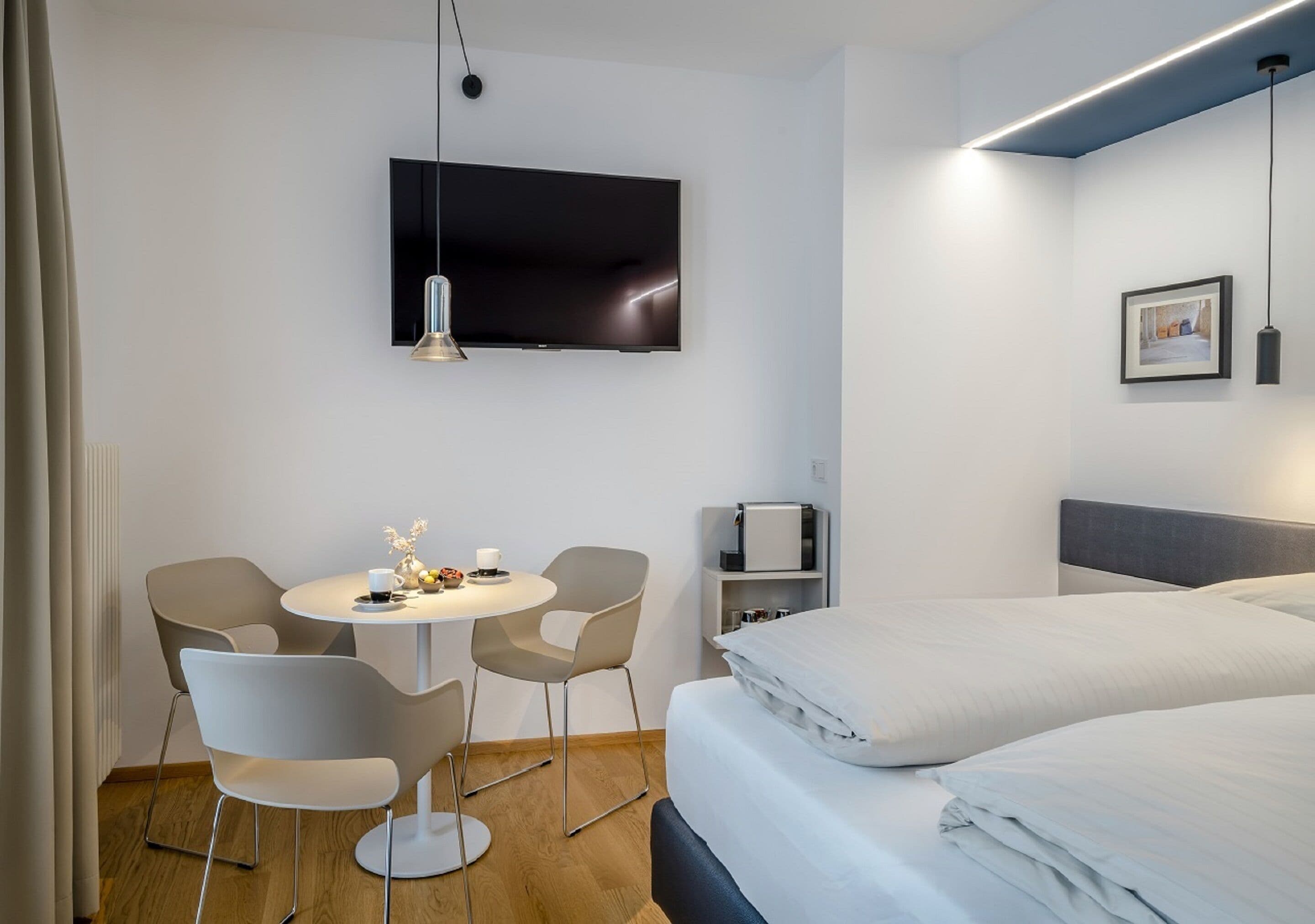 A modern hotel room with a bed, a TV mounted on the wall, a round dining table with chairs, and a small kitchenette.