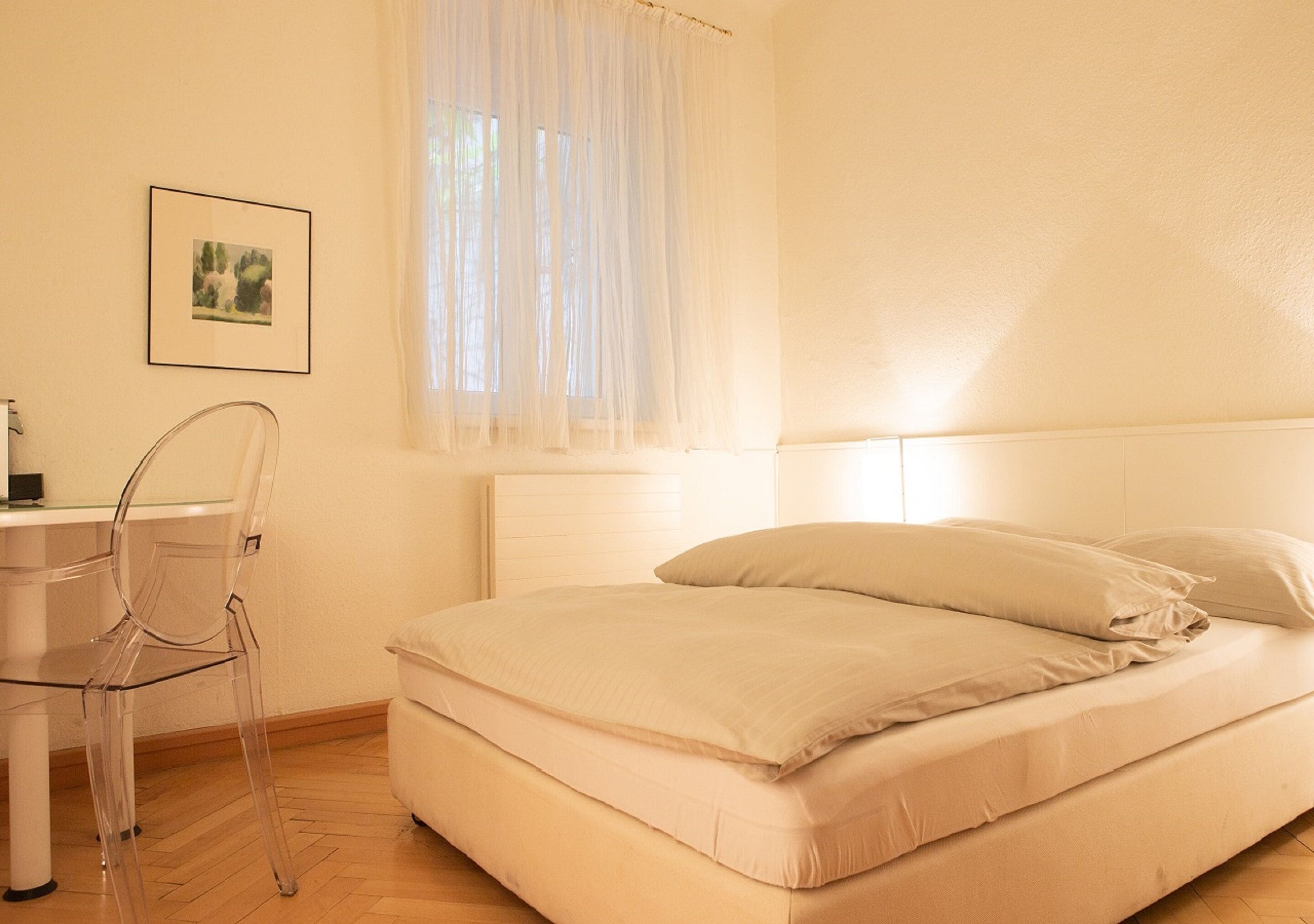 Un dormitorio con una cama, una silla, un escritorio y una ventana con cortinas de gasa.