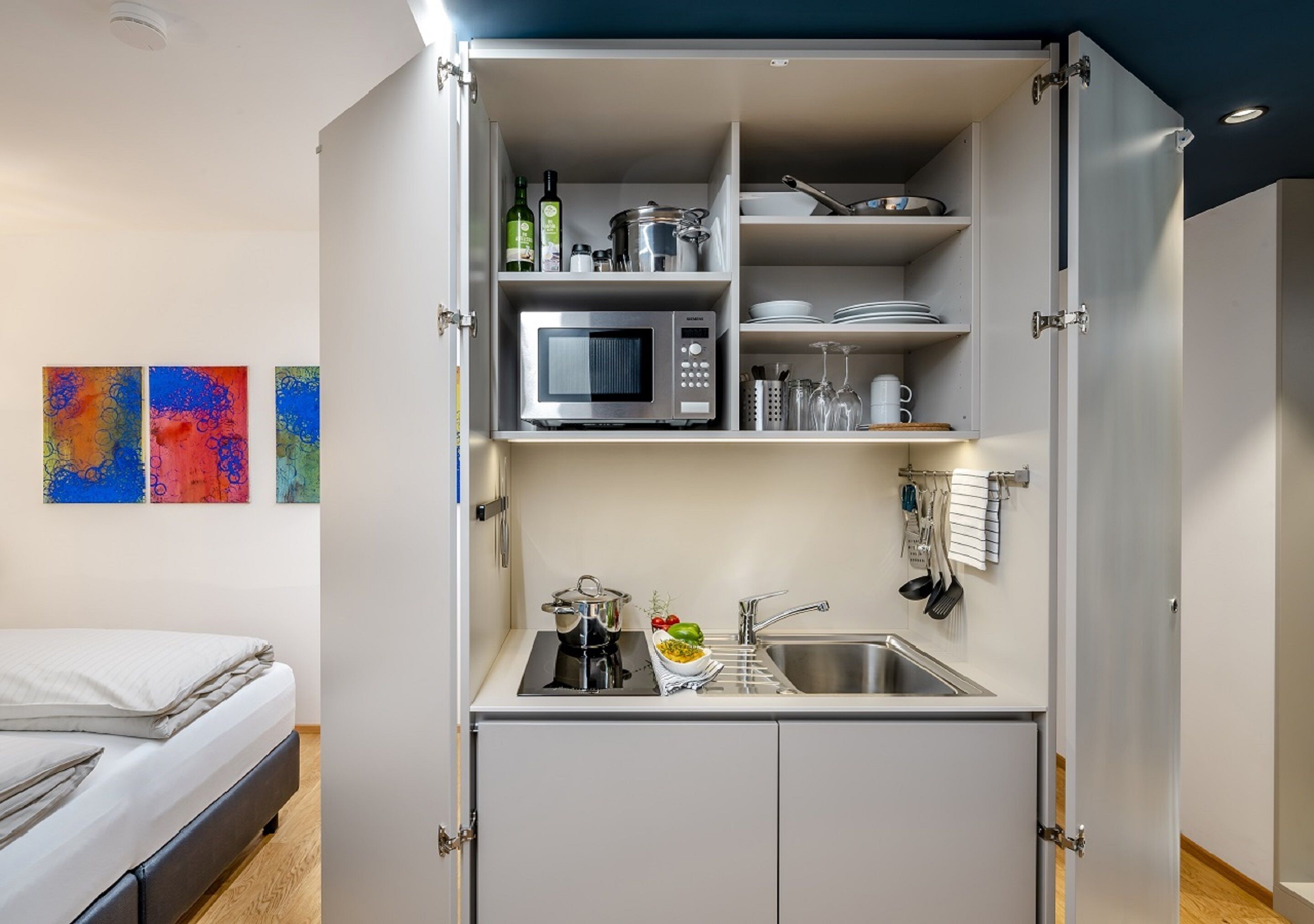 A compact kitchen with a microwave, sink, and storage cabinets. A bed is visible in the adjacent room.