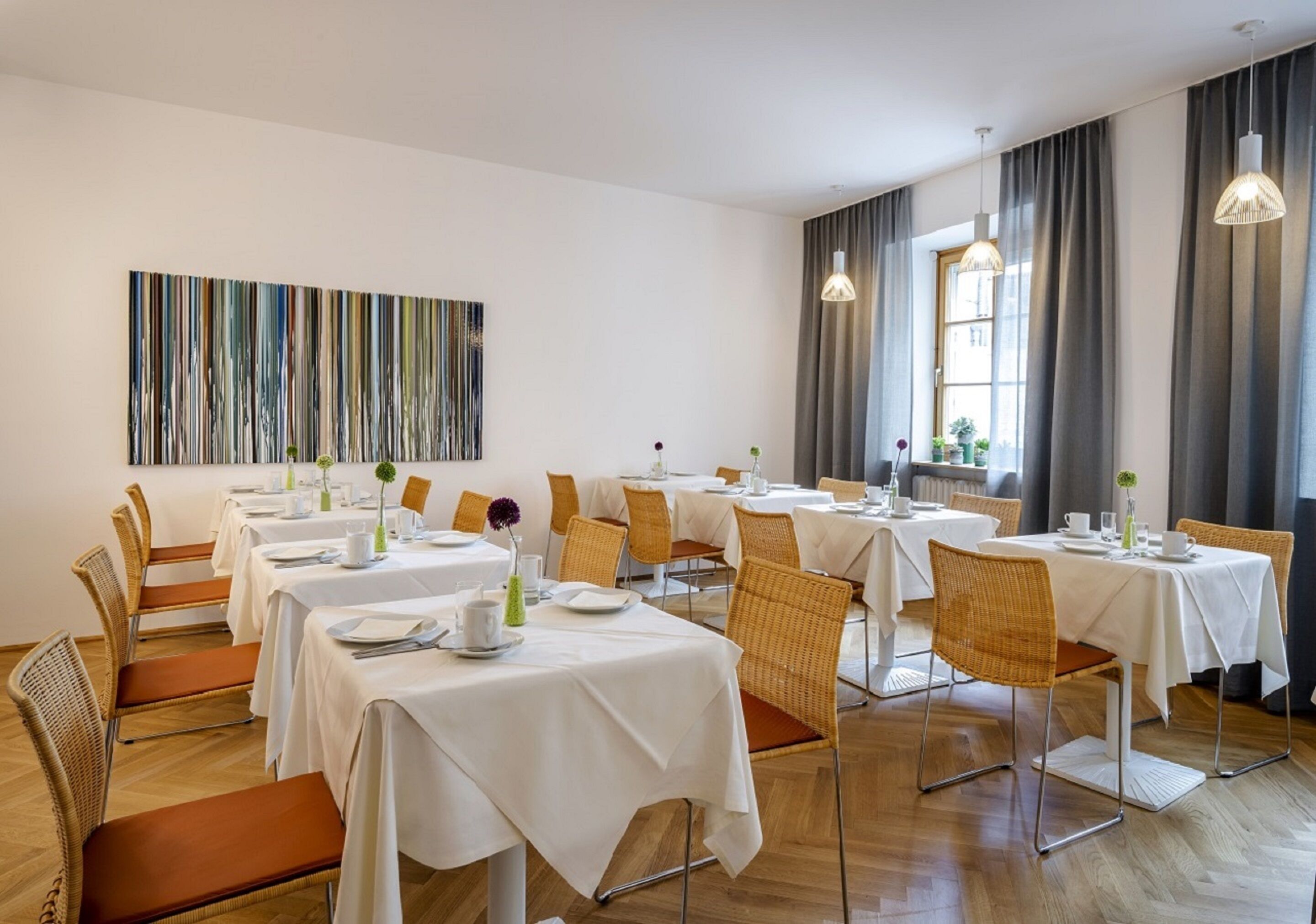 Un comedor con varias mesas y sillas, un cuadro abstracto de gran tamaño en la pared y ventanas amplias con cortinas.