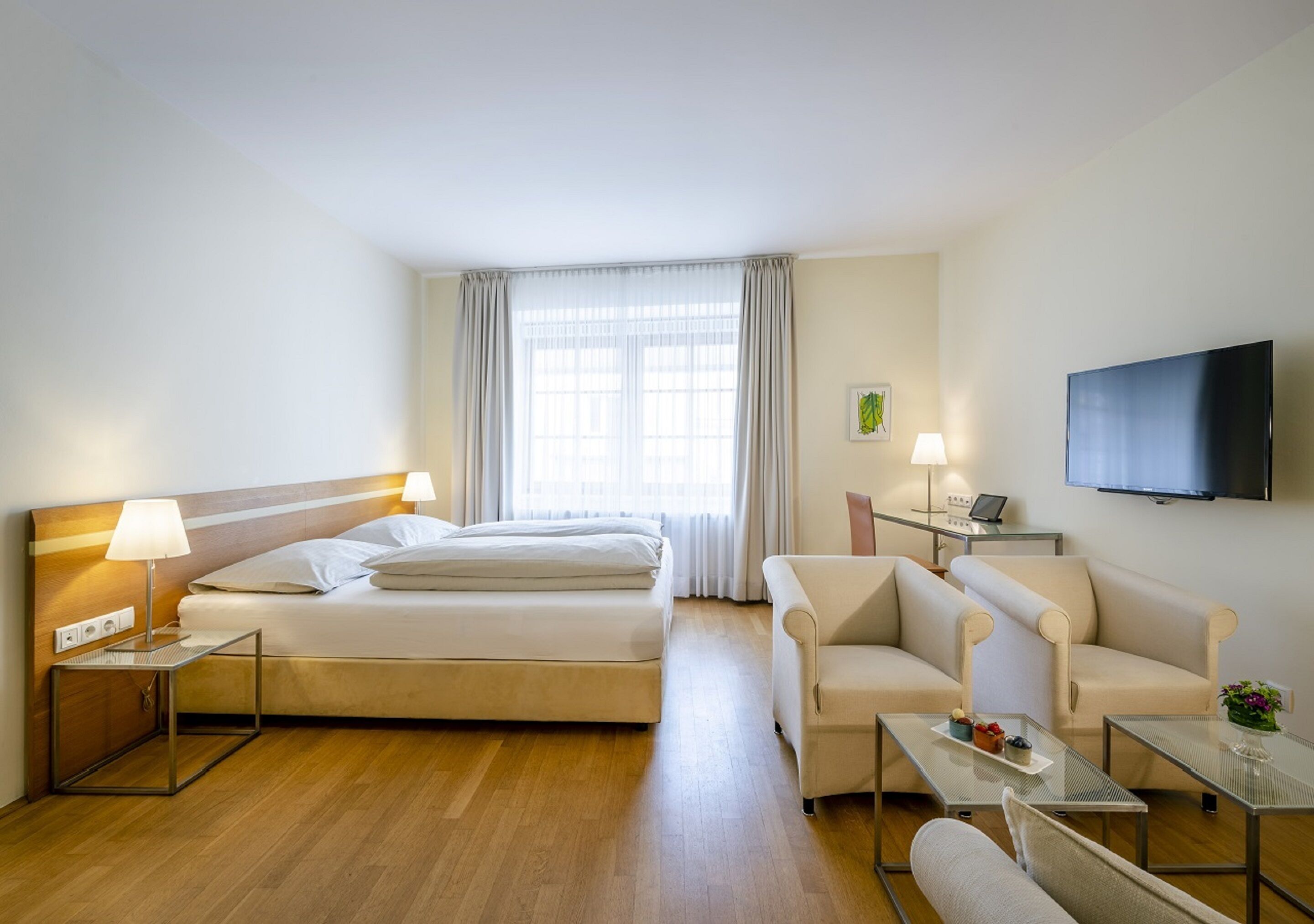 A hotel room with a bed, two bedside tables with lamps, a sofa, a TV, and a window with curtains.