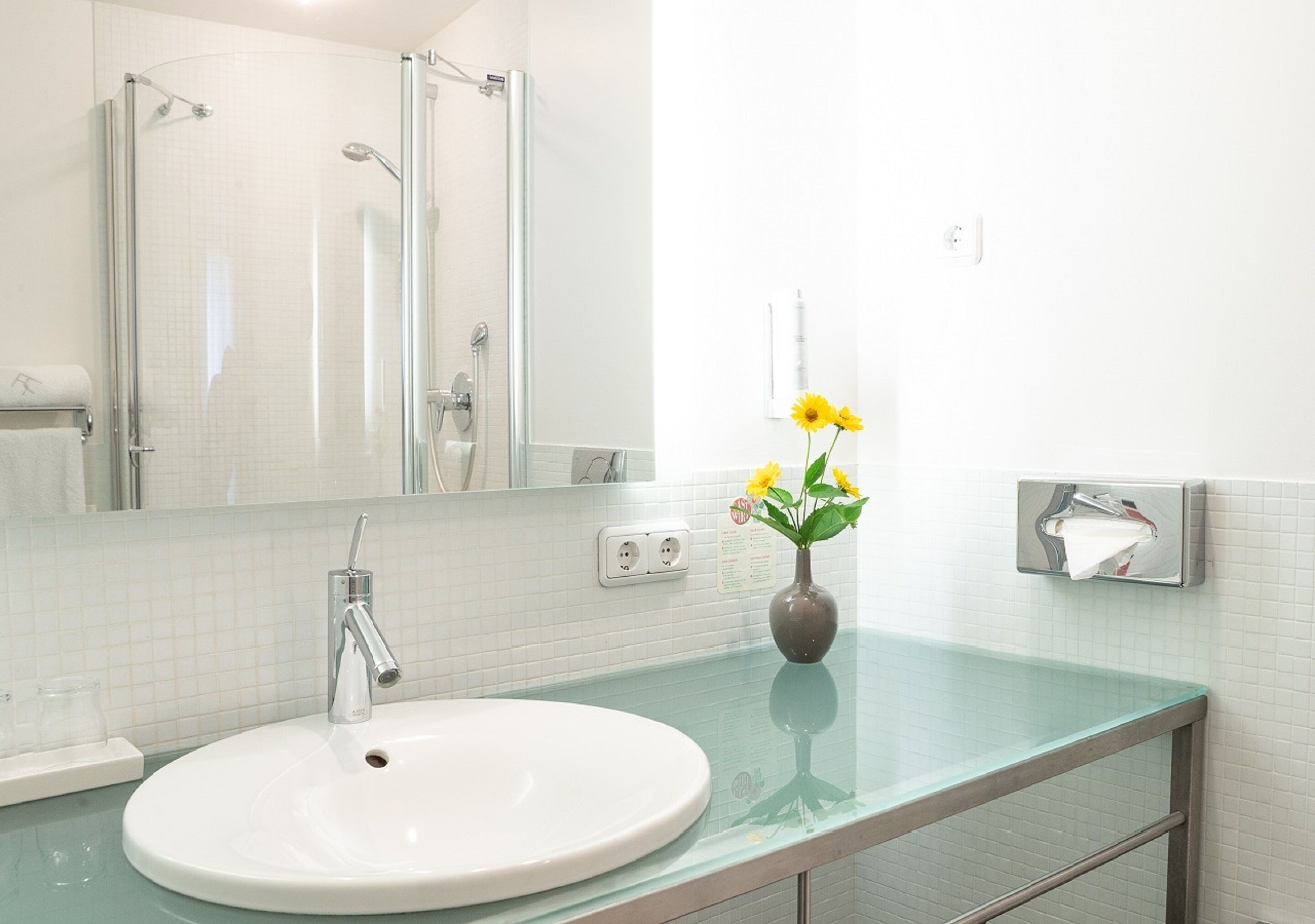Une salle de bain avec un lavabo blanc, une douche en verre et un vase rempli de fleurs jaunes posé sur le comptoir.