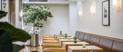 A modern interior with a row of wooden tables and benches, potted plants, and wall-mounted lighting.