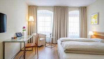 Et hotelværelse med seng, skrivebord med stol, lille bord med lampe og fjernsyn.