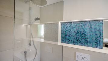 Un baño moderno con ducha envuelta en vidrio, pared con diseño de mosaico y encimera blanca.