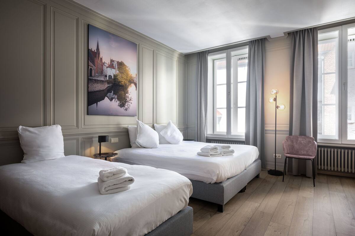 A hotel room with two beds, a large window with curtains, a floor lamp, and a painting on the wall.