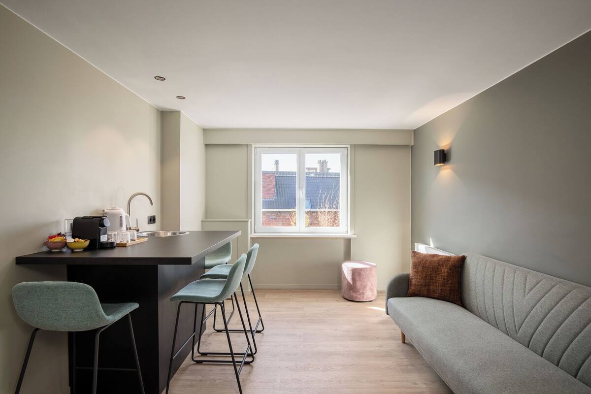 A modern kitchen with a dining area, a sofa, and a window with a city view.