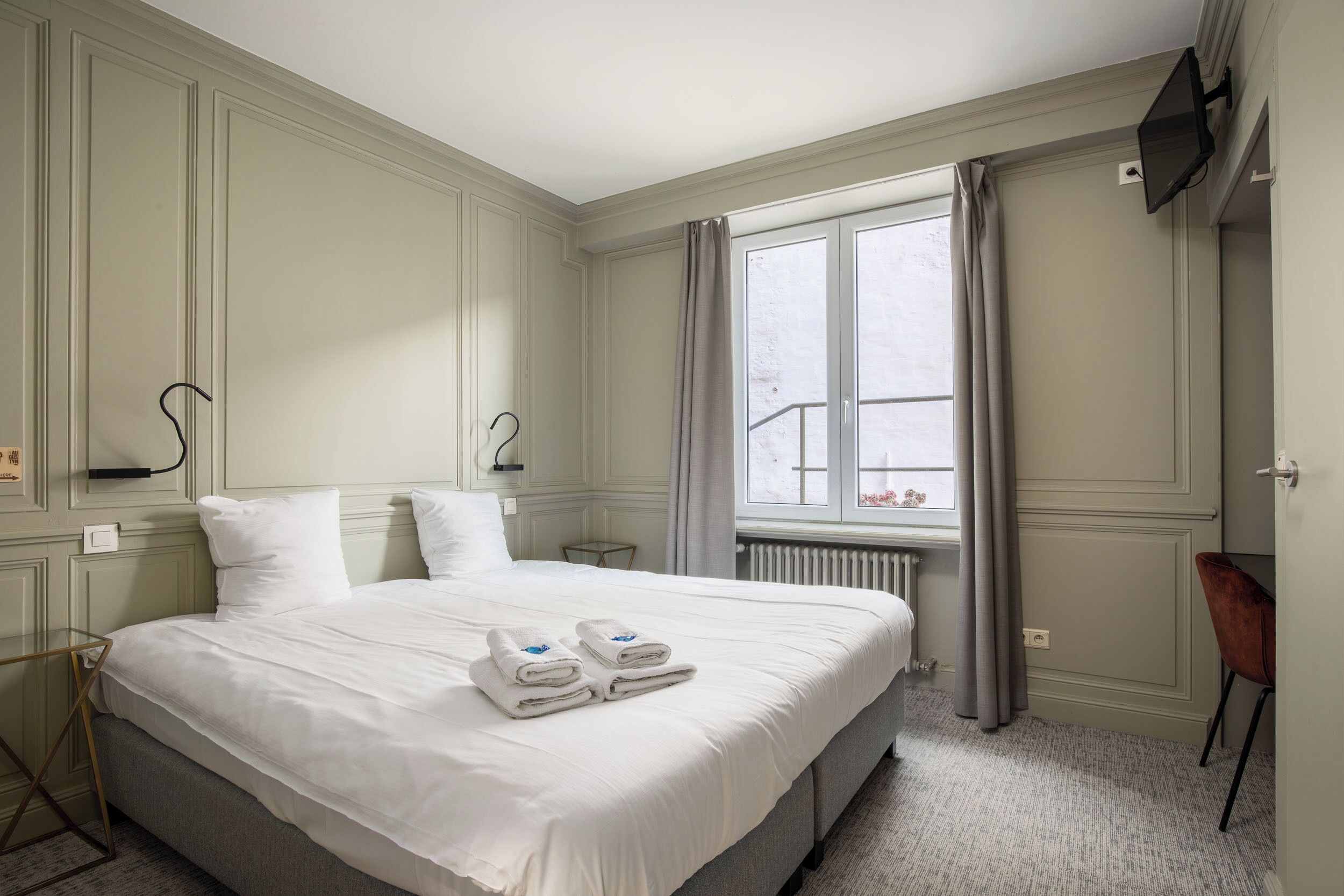 Ein Hotelzimmer mit einem großen Bett, einem Schreibtisch, einem Sessel, einem Fenster mit Vorhängen und einem an der Wand befestigten Flachbildfernseher.
