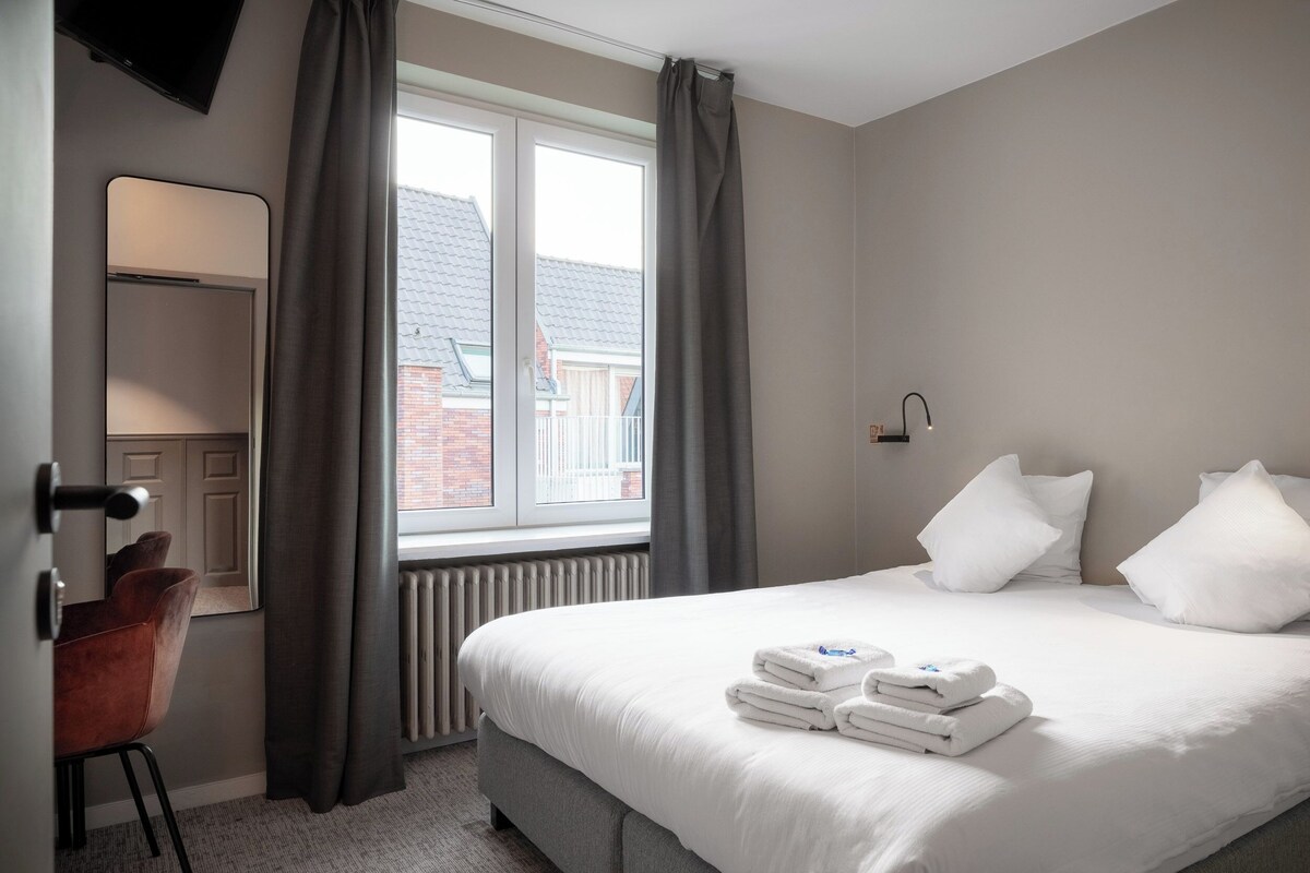 A hotel room with a bed, a desk, a chair, a window with curtains, and a radiator.