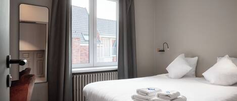 A hotel room with a bed, a desk, a chair, a window with curtains, and a radiator.
