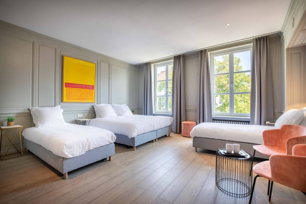 A modern hotel room with two beds, a seating area, and large windows.