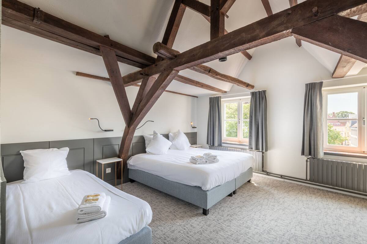 A hotel room with two beds, a wooden ceiling, a window with curtains, and a radiator.