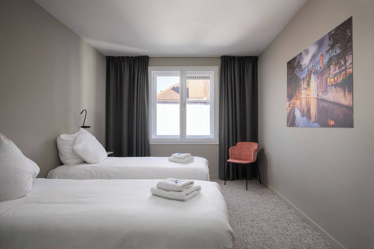 A hotel room with two beds, a window with curtains, a chair, and a painting on the wall.