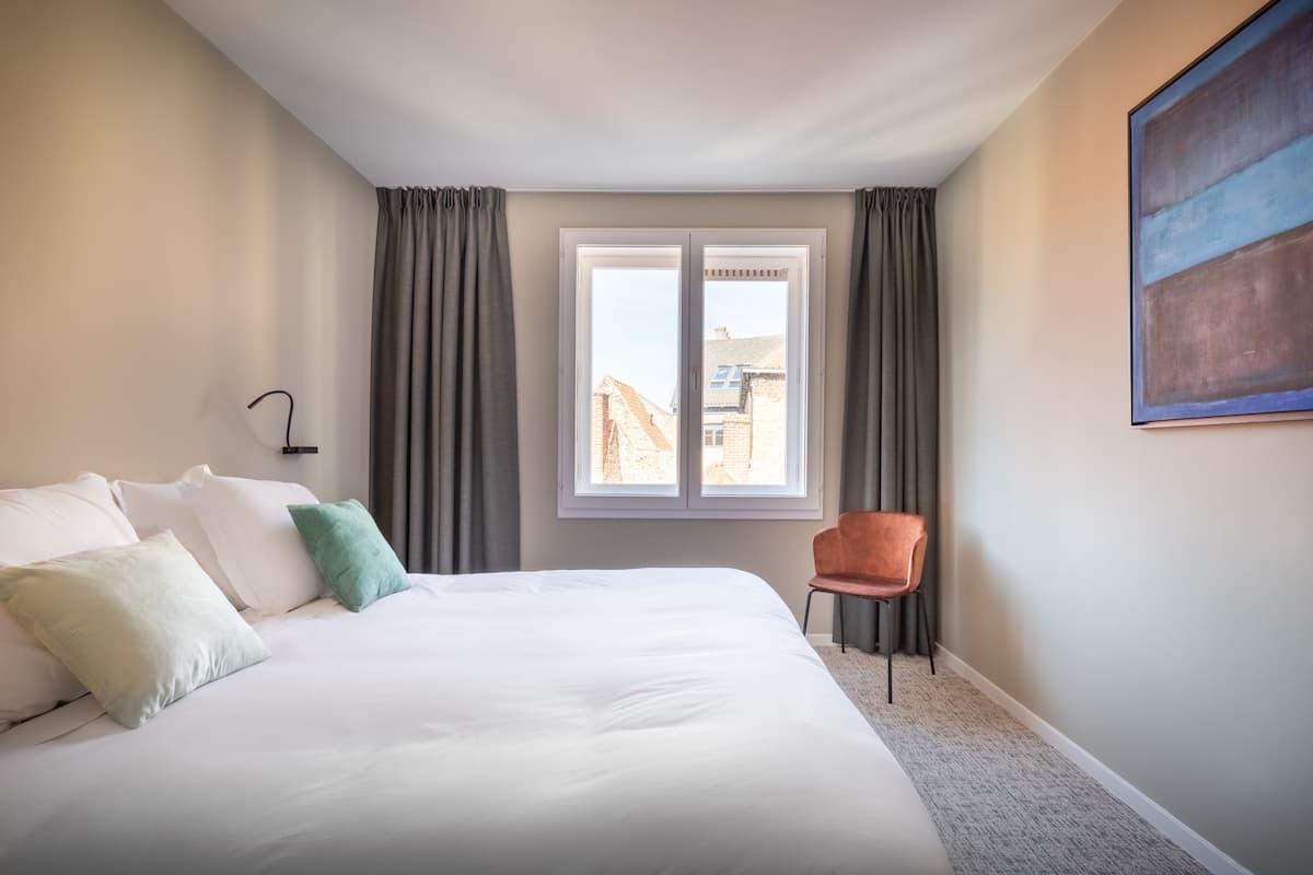 A hotel room with a large bed, a chair, a window with curtains, and a painting on the wall.