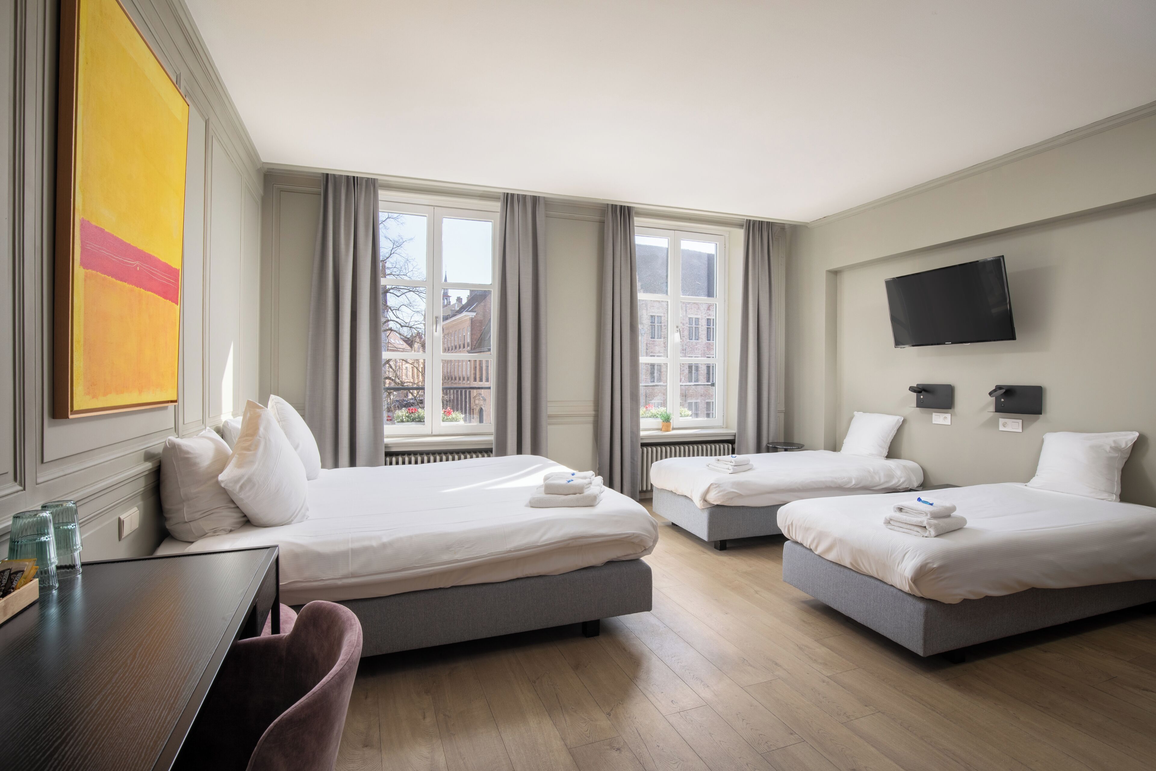 A hotel room with two beds, a desk, a TV, and large windows.