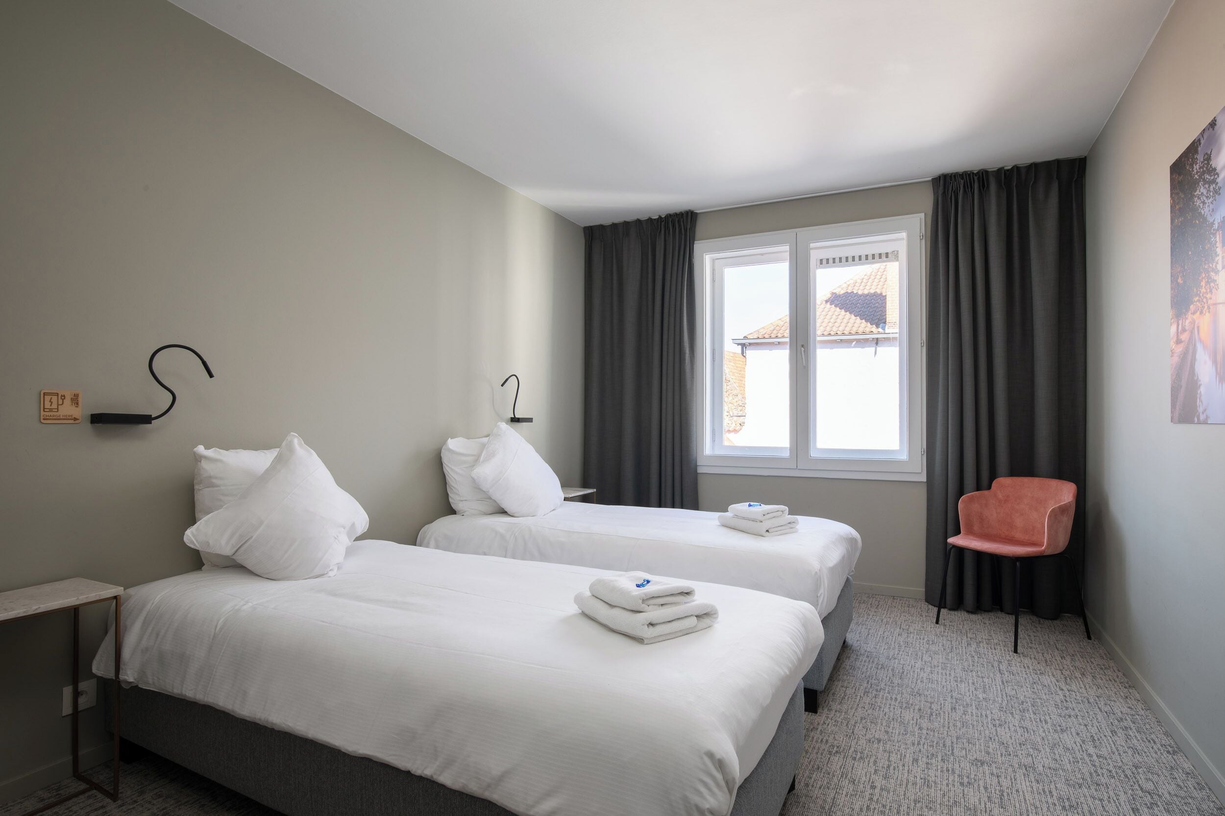 A hotel room with two beds, a desk, a chair, a window with curtains, and a wall-mounted light fixture.
