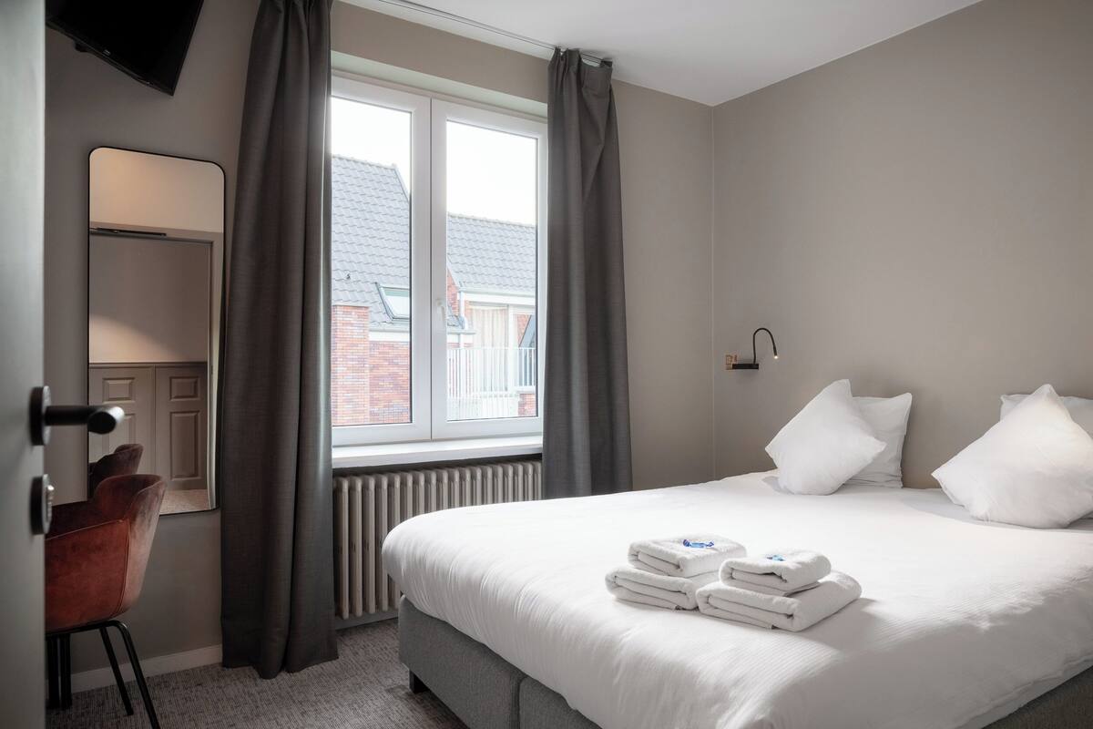 A hotel room with a bed, a desk, a chair, a window with curtains, and a radiator.