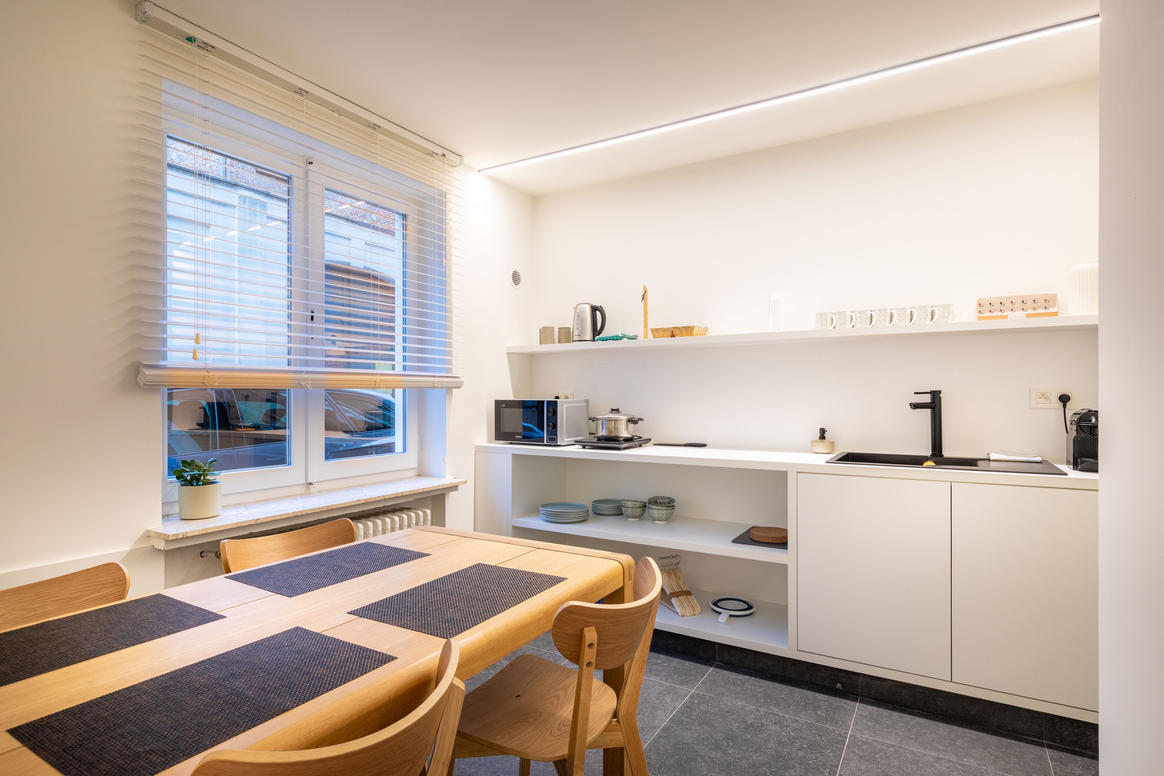 Une cuisine moderne avec des meubles blancs, un four à micro-ondes encastré et un évier noir. On y trouve une table à manger avec des chaises et une fenêtre équipée de stores.