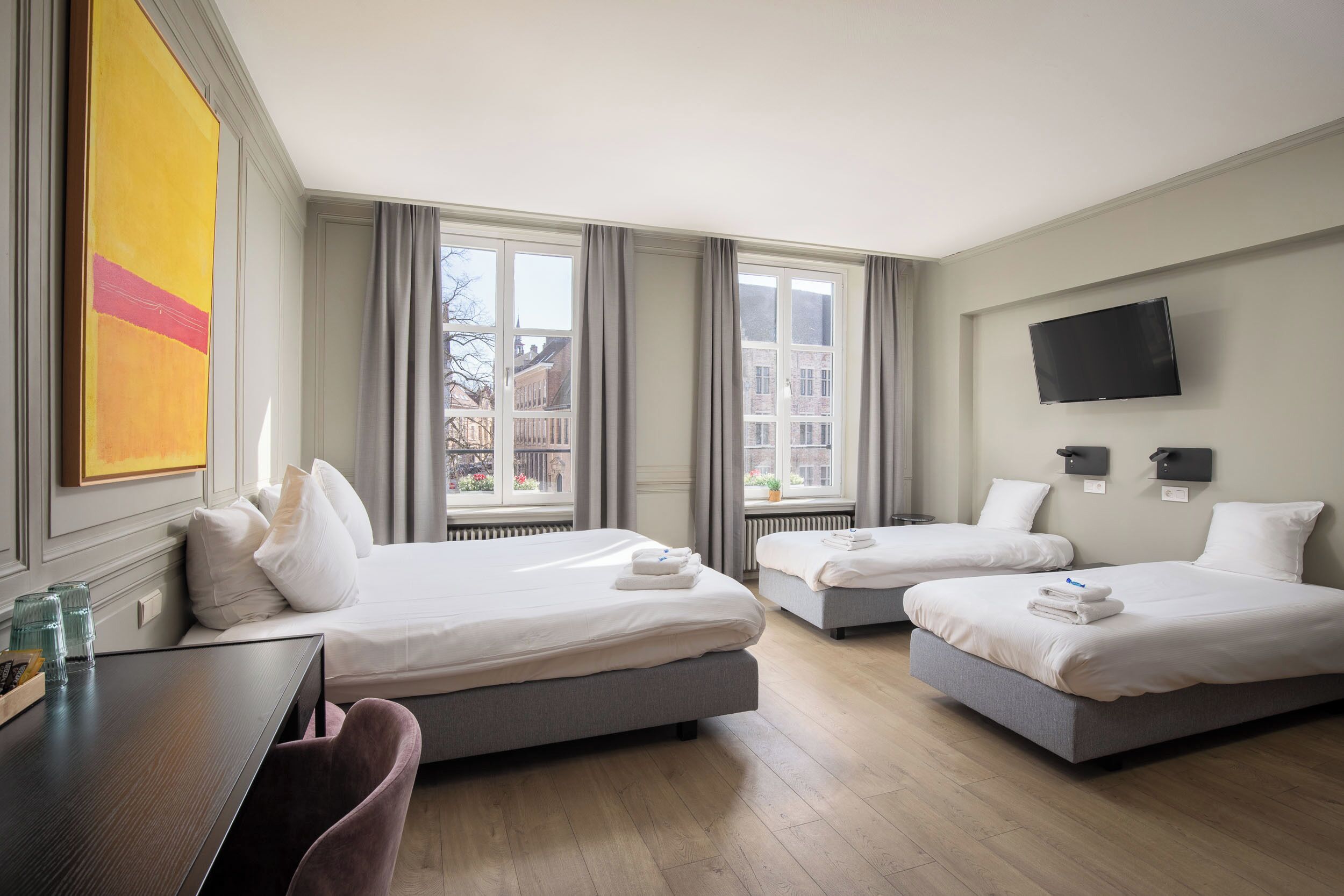 Et hotelværelse med to senge, et skrivebord, et fjernsyn og store vinduer.