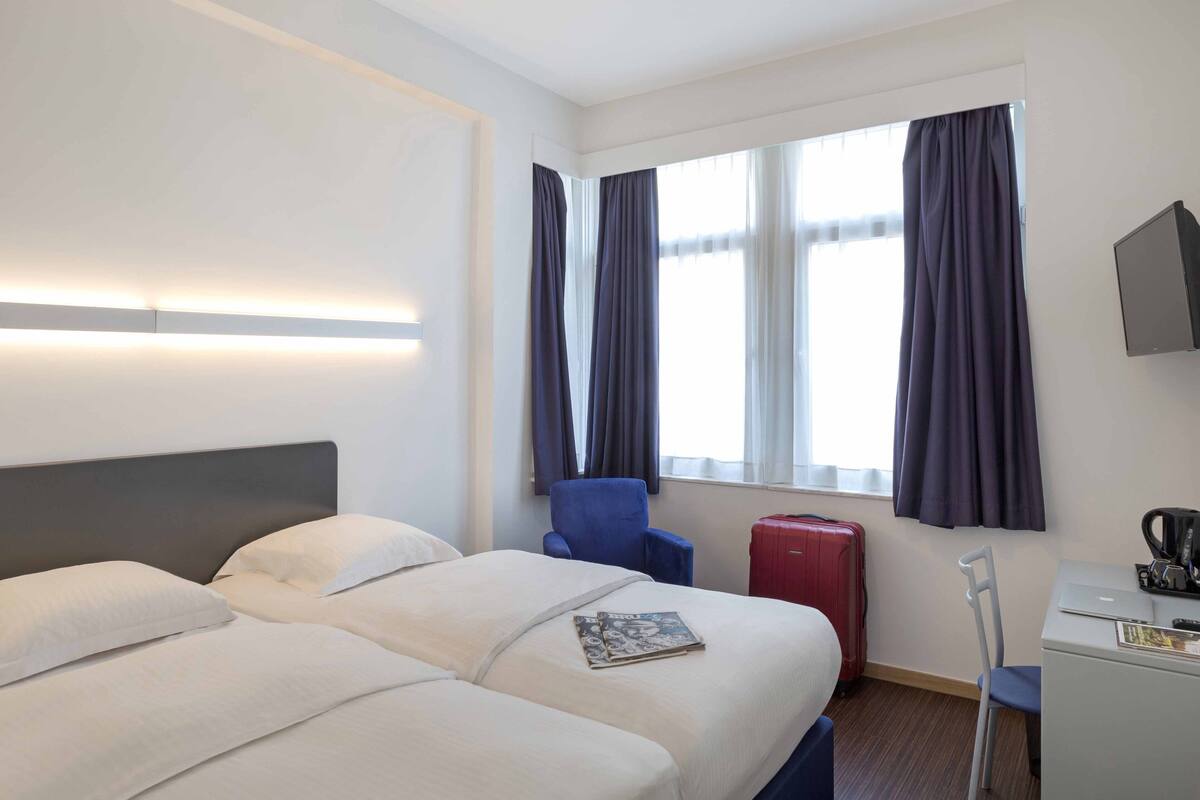 Habitación de hotel con dos camas, un escritorio, una silla, una maleta roja y un ventanal con cortinas.