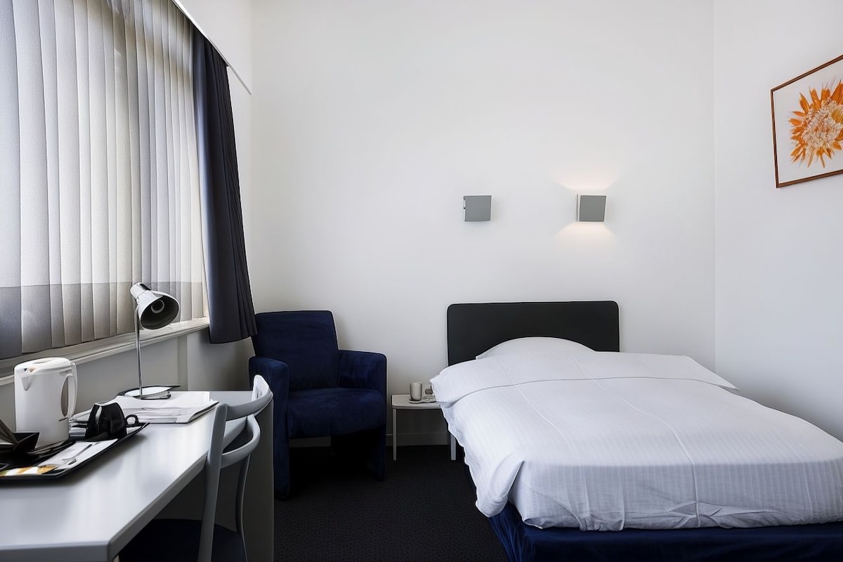 A hotel room with a bed, a desk with a chair, a nightstand, a lamp, and a window with blinds.